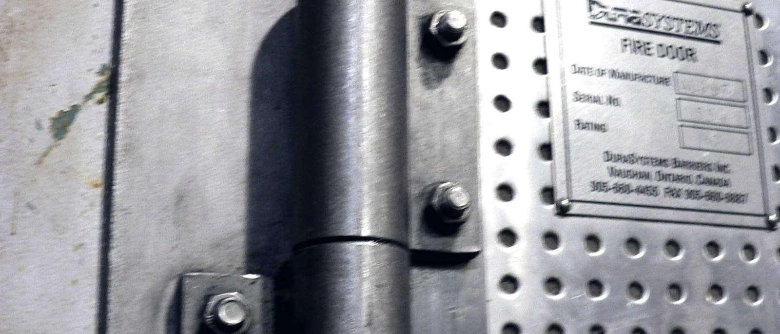 Close-up of a heavy steel butt hinge on a perforated fire-door leaf beside a stamped certification plate