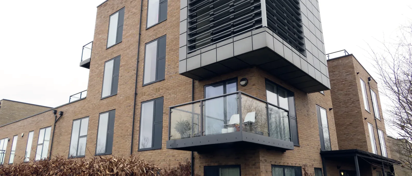 An upward view of a modern brick apartment block corner showing a glazed balcony and a smoke ventilation louvre unit at roof level