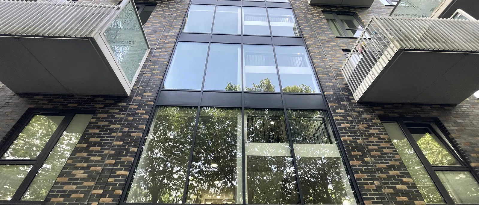 A dramatic upward view of a glazed stair and lift tower with glass-box balconies projecting from a brick facade against a blue sky