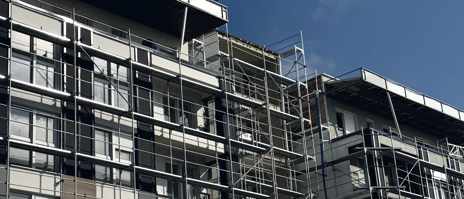 Scaffolding erected against the balconied facade of a mid-rise residential block under a blue sky