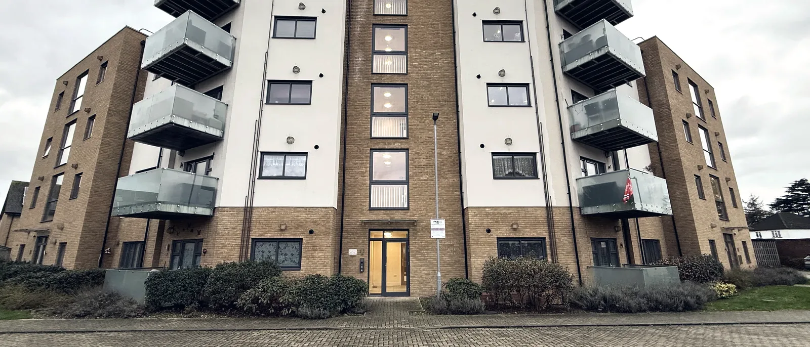 A modern white-clad residential apartment block with a grid of windows and balconies, seen from the entrance path with hedges in the foreground