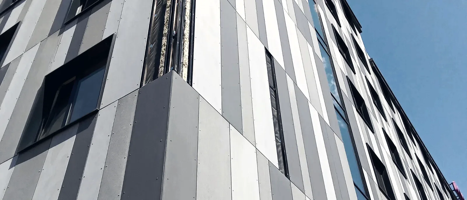 A dramatic upward view of a grey and charcoal panelised rainscreen cladding facade against a blue sky with clouds