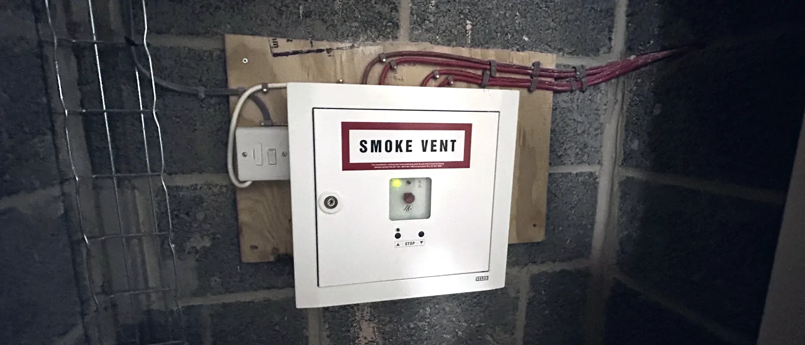 A white 'Smoke Vent' control panel with an indicator light, mounted on a plywood backing board over a breeze-block wall with exposed cabling