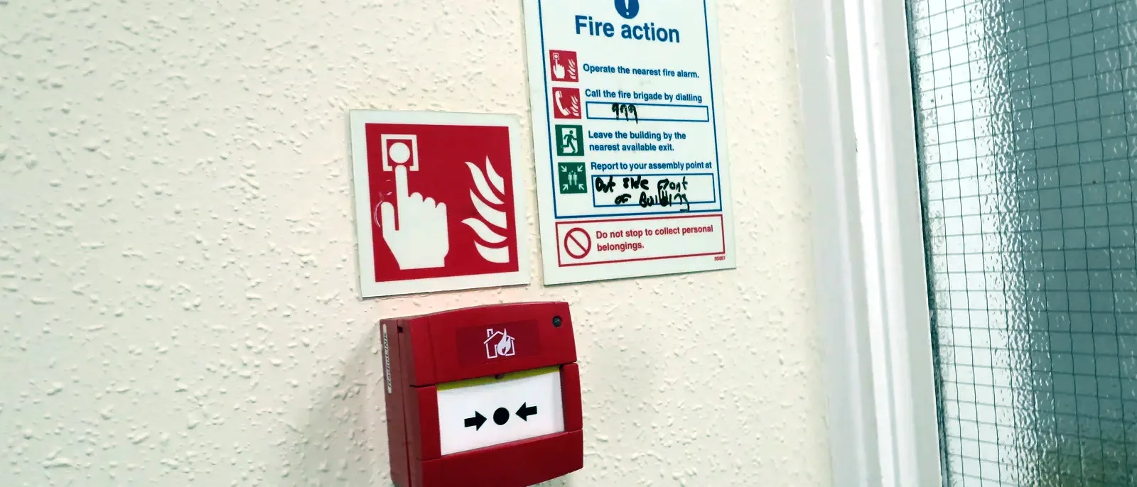 A red 'Fire action' instruction notice mounted above a red manual call point on a textured wall, beside a wired-glass door panel