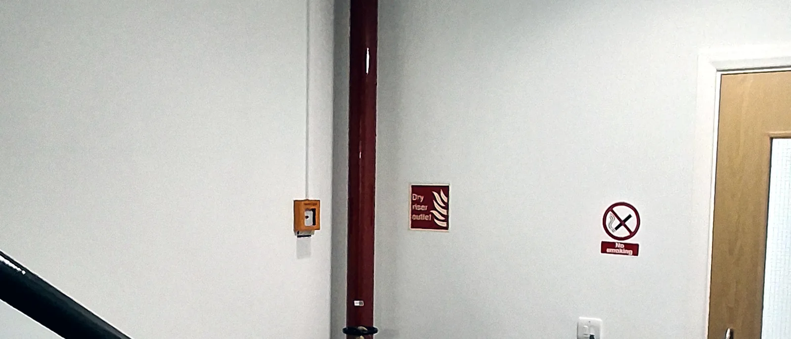 A stairwell landing with red dry riser pipework running up the wall beside a black metal handrail