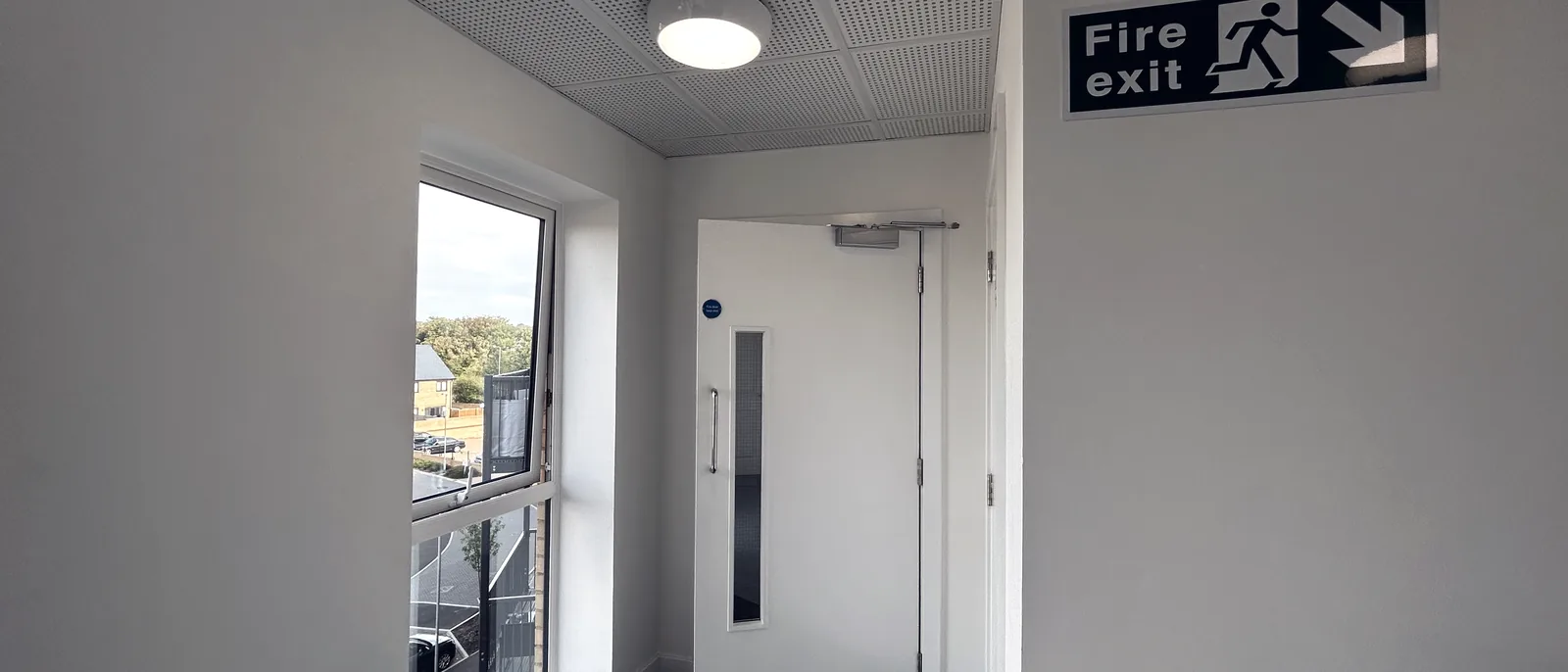 A corridor with a glazed fire door fitted with an overhead closer, and an illuminated fire exit sign on the wall alongside