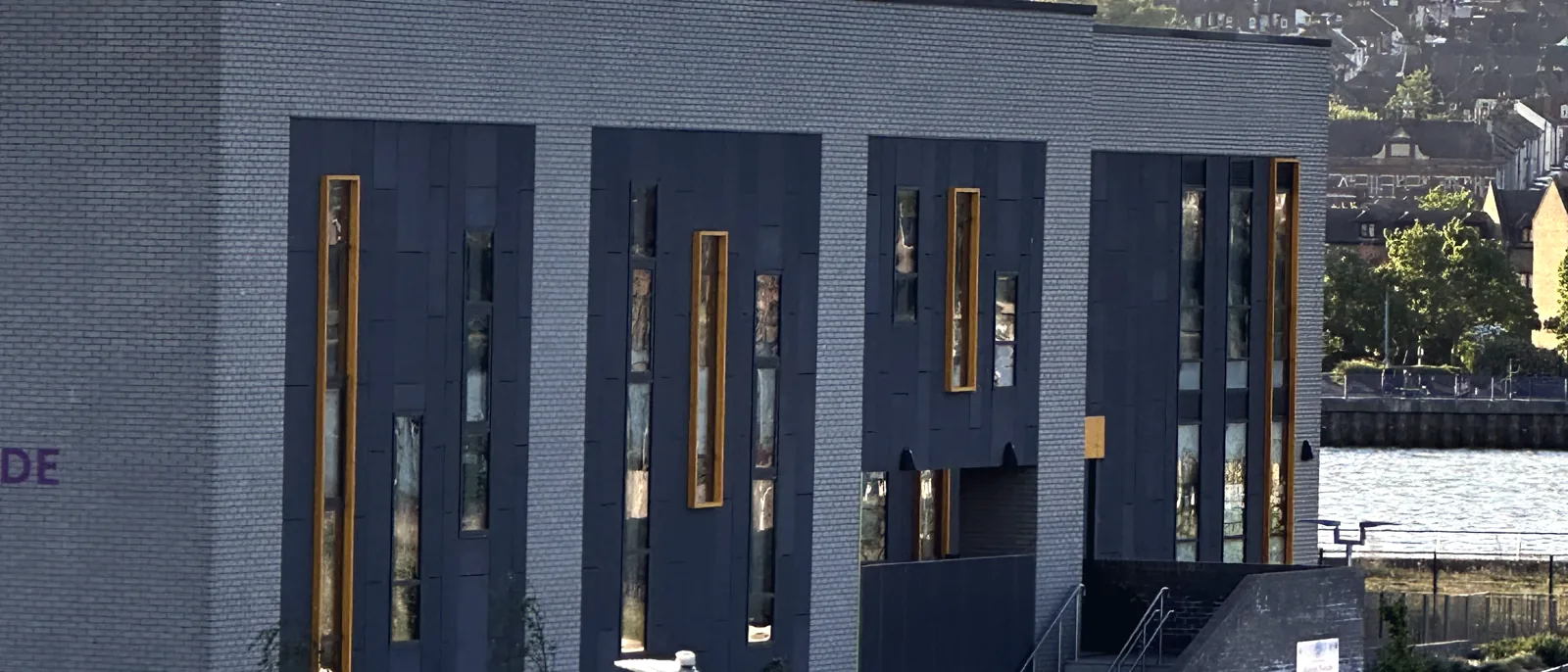 A modern grey-brick school building with tall narrow windows above a landscaped riverside path