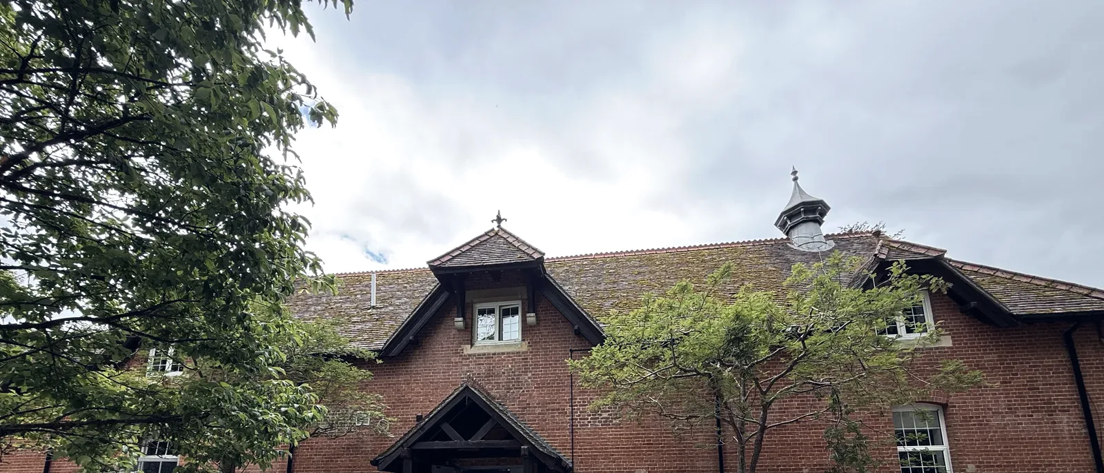 A red-brick period building with a tiled roof, dormer windows and a timber entrance porch, seen through trees