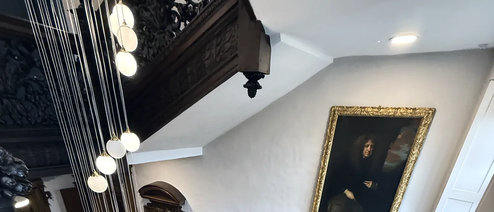 A carved dark wood ceiling beam and hanging globe pendant lights beside a staircase landing, with a gilt-framed portrait on a pale wall