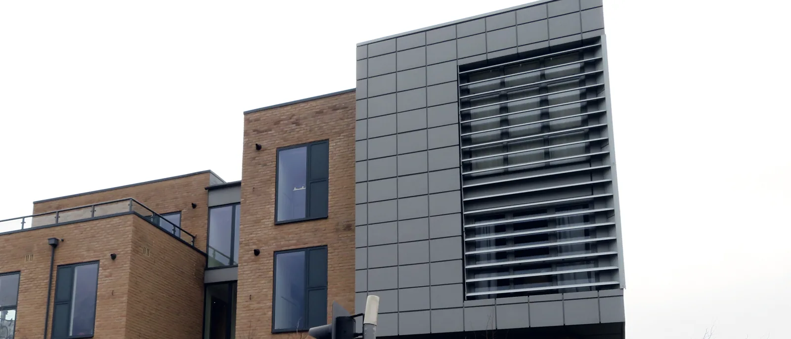 The corner of a building where dark grey rainscreen cladding panels meet a brick facade with windows