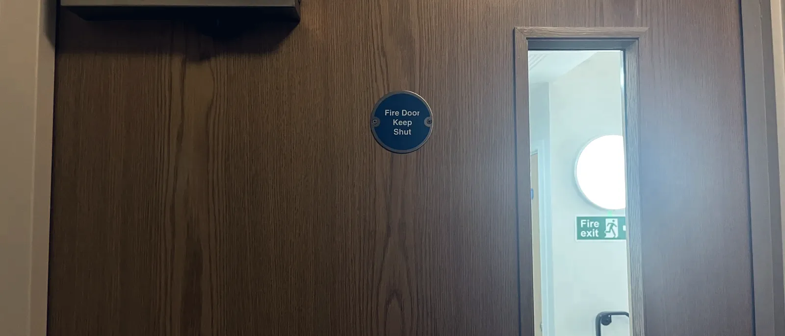 A timber fire door with an overhead closer, a wire-mesh vision panel and a keep-shut disc, a fire exit sign visible in the corridor beyond